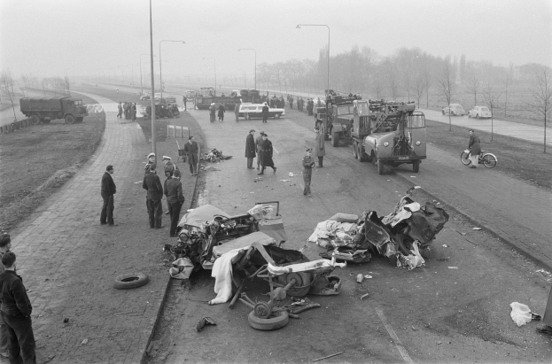 Schwarz-weiß-Szene eines Autounfalls am Rande einer Straße mit mehreren Fahrzeugen und einer Gruppe von Menschen drumherum, Lichtmasten, Bäume und Himmel im Hintergrund.