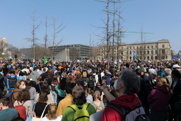 Eine große Menschenmenge bei einer Klimaprotestveranstaltung vor einem Gebäude mit Bäumen und einem klaren Himmel im Hintergrund.