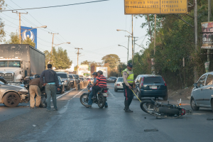 Eine Gruppe von Menschen steht um ein verunglücktes Motorrad auf der Straße mit mehreren Fahrzeugen, darunter ein Lastwagen, und einem Hintergrund aus Bäumen, Pfählen, Lampen, Schildern und Himmel.