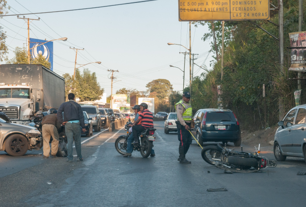Eine Gruppe von Menschen steht um ein verunglücktes Motorrad auf der Straße mit mehreren Fahrzeugen, darunter ein Lastwagen, und einem Hintergrund aus Bäumen, Pfählen, Lampen, Schildern und Himmel.