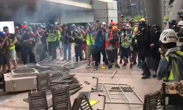 Eine Gruppe von Menschen in Helmen und Jacken steht vor einem rauchenden Gebäude, einige halten Kameras, mit verstreuten Metallobjekten und Stadtgebäuden im Hintergrund.