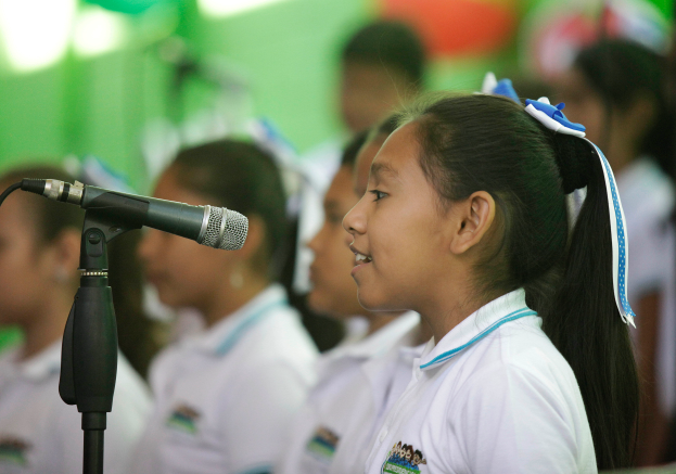 Eine Gruppe junger Mädchen in weißen T-Shirts steht vor einem Mikrofon, wobei das Mädchen in der Vordergrund den Fokus bildet.