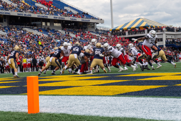 Fußballspieler mit Helmen konkurrieren auf einem Rasenfeld, während Zuschauer auf Tribünen unter einem bewölkten Himmel sitzen.