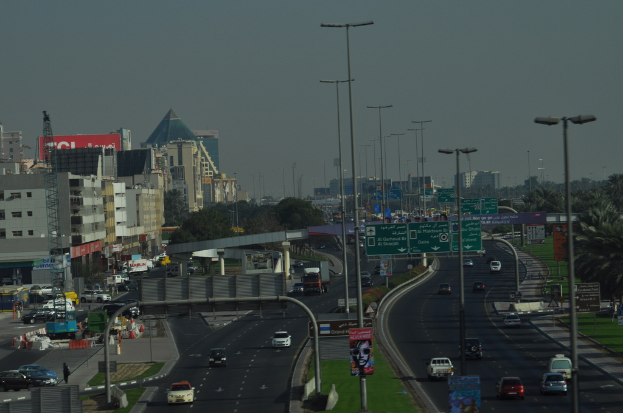 Luftaufnahme einer Stadt mit Gebäuden, Türmen, Kraftfahrzeugen, Werbetafeln, Schildern, Straßen, Bäumen, Straßenlaternen, Menschen, Verkehrsleitkegeln und Informationsschildern unter einem klaren Himmel.