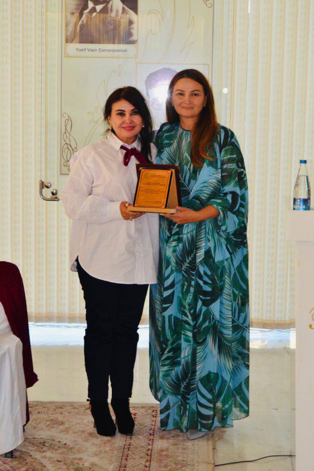 Zwei Frauen stehen nebeneinander und lächeln auf einer Preisverleihung; die Frau links hält eine Auszeichnung, während die Frau rechts neben einem Podium mit einer Wasserflasche steht, mit Vorhängen und einem Foto im Hintergrund.