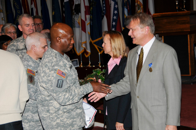 Eine Gruppe von Menschen, die stehen, mit zwei Personen, die sich die Hände schütteln, im Vordergrund, Fahnen an Stangen, ein Podium mit einem Mikrofon und eine Topfpflanze in der Nähe.