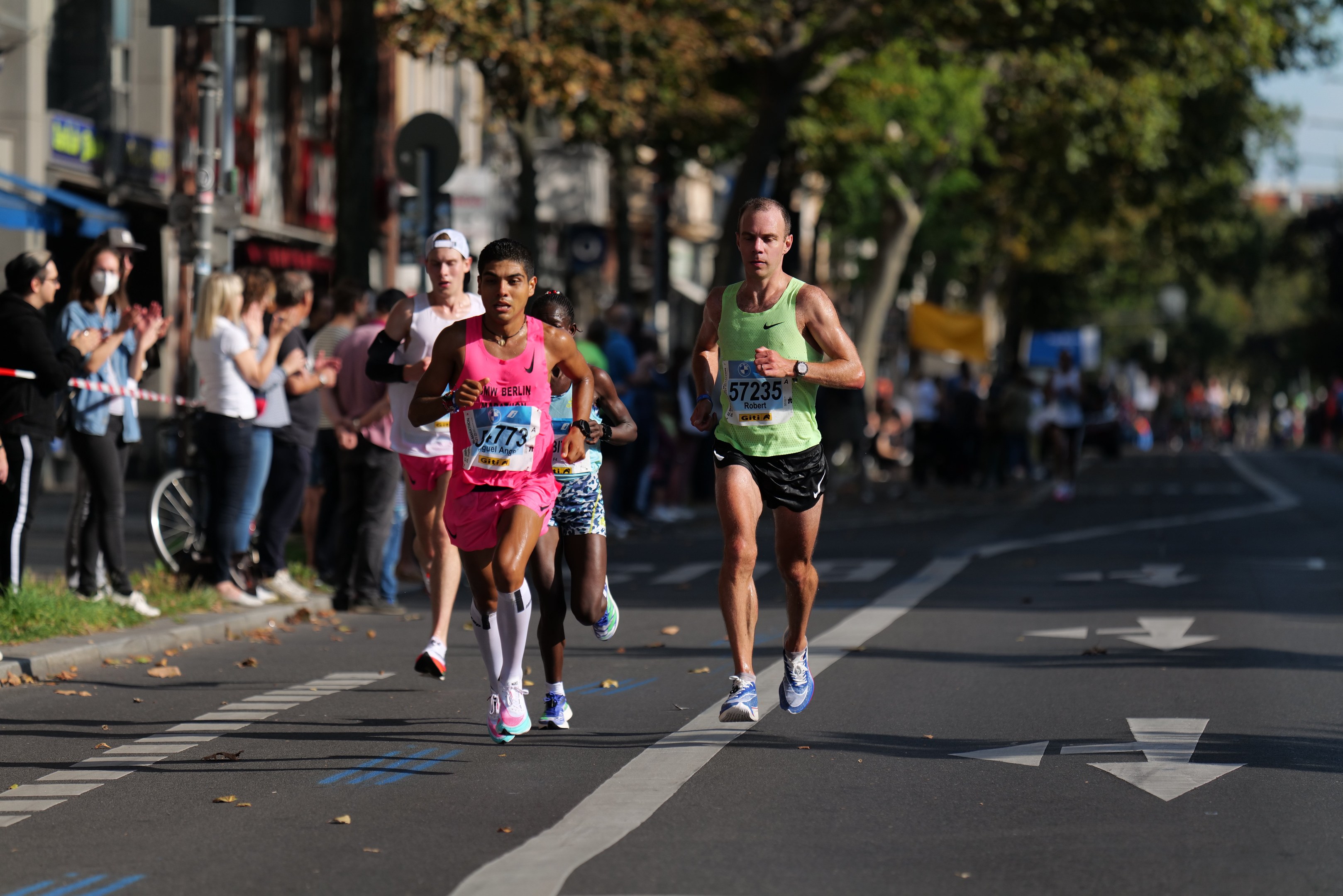 Gruppe von Läufern bei einem Marathon auf einer Stadtstraße, mit Zuschauern und städtischen Elementen im Hintergrund.