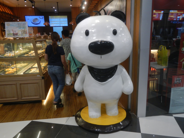 Eine Riesengänse-Statue steht in einem Laden, umgeben von Einkäufern, mit einer Auslage mit Lebensmitteln links und Fernsehern an der Decke im Hintergrund.