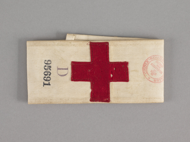 Ein rotes Kreuz-Ersthilfepaket in einer Stofftasche mit Text und einem Stempel, das auf einer Oberfläche liegt.