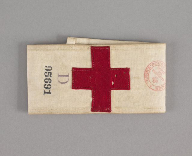 Ein rotes Kreuz-Ersthilfepaket in einer Stofftasche mit Text und einem Stempel, das auf einer Oberfläche liegt.