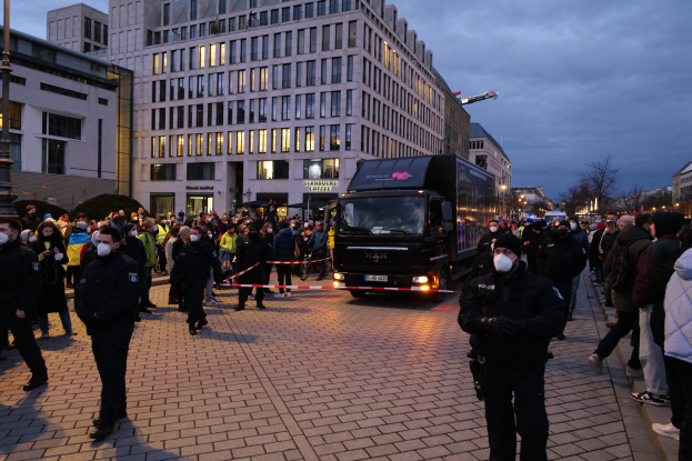 Eine Gruppe von Menschen steht vor einem Lastwagen auf einer Straße, umgeben von Gebäuden, Laternenmösten, Bäumen und einem bewölktem Himmel, wobei einige Mötzen und Masken tragen und ein Band mit einem Pfahl im Vordergrund.