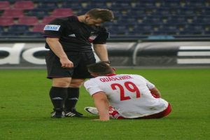 Ein Fussballspieler in Sportbekleidung sitzt neben einem Schiedsrichter auf dem Boden in einem Stadion Setting, mit Tafeln und Stühlen im Hintergrund.