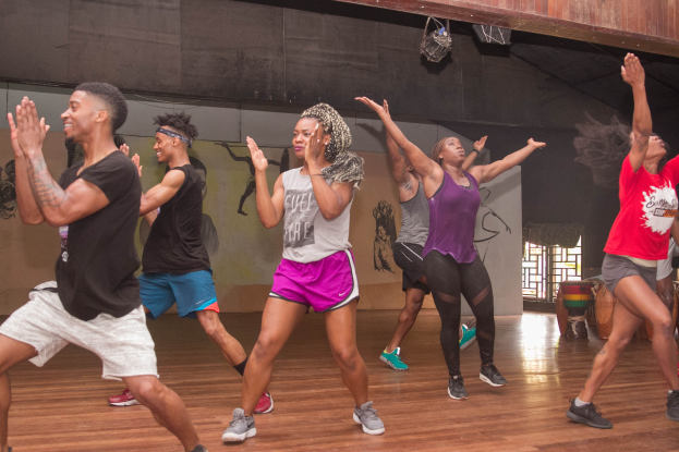 Eine Gruppe von Menschen, die in einem Studio mit einer bemalten Wand, einem Fenster und Deckenbeleuchtung tanzen, wahrscheinlich an einem Zumba-Kurs teilnehmen.