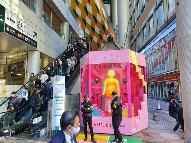 Eine Gruppe von Menschen steht vor einem Gebäude mit Glaswänden und Texttafeln, einige tragen Masken, während eine Rolltreppe mit Fahrgästen zu sehen ist; ein pinker Kasten in der Mitte zeigt Schaufensterpuppen und trägt den Text "Netflix's new Netflix Booth in Tokyo."