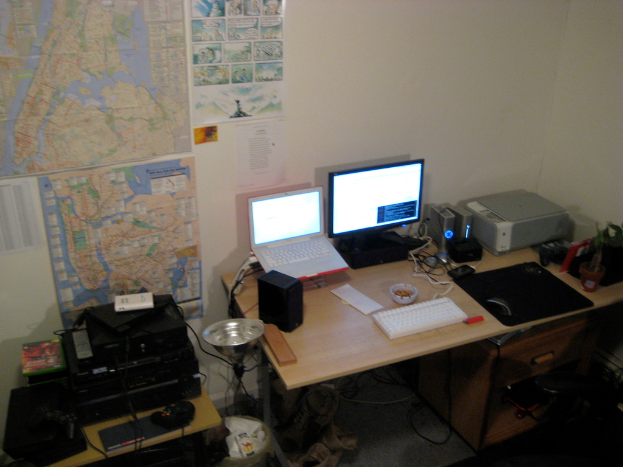 Ein Raum mit einem Tisch, auf dem ein Laptop, Lautsprecher, Monitor, Tastatur, Ball, Maus, Scanner und Papiere stehen, und einem weiteren Tisch mit Elektronikgeräten im Hintergrund, mit Charts und Postern an der Wand dahinter.