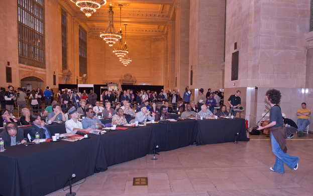 Ein Konzertbild mit einer Menschenmenge, einem Mann, der auf der rechten Seite Gitarre spielt und singt, einer Wand und Fenstern im Hintergrund, Lichtern oben und einem Boden unten.