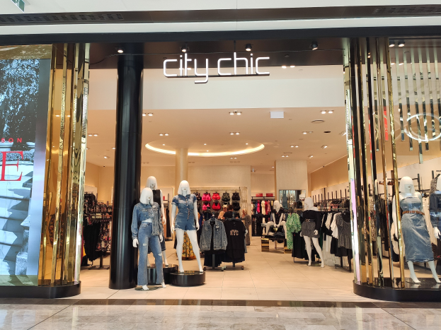 Ein stylischer Stadtmode-Laden in einem Einkaufszentrum mit Schaufensterpuppen in der neuesten Mode, Kleidung an Bügeln, einer Säule, einem Schild, Deckenlampen und einem gefliesten Boden.