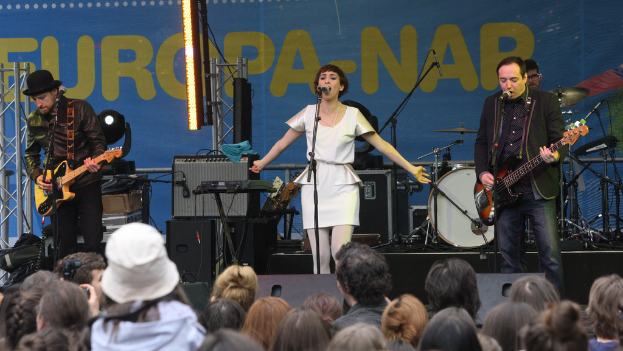 Eine Bühne mit vier Musikern: eine Frau in einem weißen Kleid singt in der Mitte, zwei Männer spielen Gitarren auf der linken und rechten Seite und eine Menge ist unten zu sehen, mit einer Wand und Lautsprechern im Hintergrund.