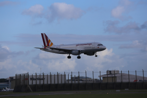 Germanwings Airbus A320-200 beim Start von Frankfurt Airport mit Gras darunter, einem Zaun im Vordergrund, Gebäuden und Bäumen im Hintergrund und einer bewölkten Himmel