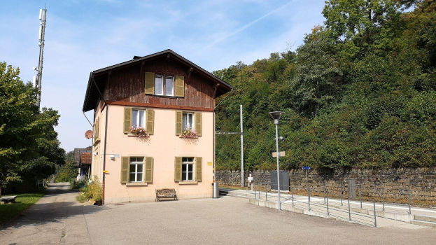 Ferienhaus zum Verkauf in Baden-Württemberg, Deutschland, mit einem Gebäude mit zahlreichen Fenstern, einer vorderen Mauer, umliegenden Straßen-Elementen, Grünflächen und einem bewölkten Himmel.