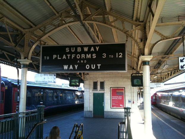 Zwei Züge stehen neben einem Bahnsteig mit zwei Bildschirmen und mehreren Schildern, die von der Decke hängen, eine Person steht in der Nähe der Treppe, und eine Tür ist in der Wand.