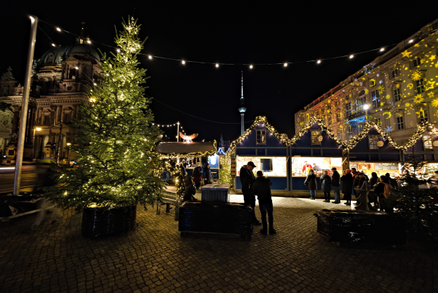 Ein lebendiger Weihnachtsmarkt in Berlin, Deutschland, zeigt einen großen, hell erleuchteten Weihnachtsbaum in der Mitte, umgeben von Menschen und Gebäuden mit Fenstern unter einem dunklen Himmel.