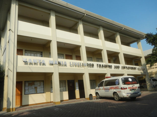Außenansicht des Santa Maria Lebensmittel-Trainings- und Beschäftigungszentrums mit Fenstern, Türen und Beschriftung, umgeben von Bäumen und parkenden Fahrzeugen unter einem klaren blauen Himmel.