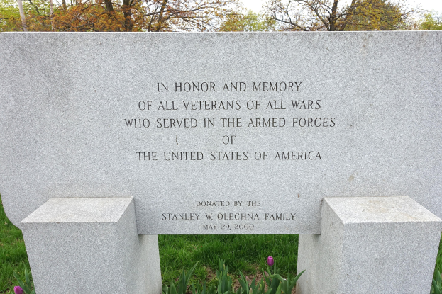 Ein Gedenkstein mit der Inschrift "In Ehren und Andenken aller Veteranen aller Kriege, die in den Streitkräften der Vereinigten Staaten von Amerika dienten", umgeben von Pflanzen, Blumen, Gras, Bäumen und einem bewölkten Himmel.