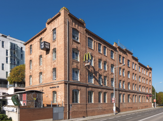 Ein großes McDonald's-Restaurantgebäude aus Backsteinen an einer Straßenecke in Berlin, Deutschland, mit umgebender Straßeninfrastruktur, Fahrzeugen, Grünflächen und einem bewölkten Himmel.