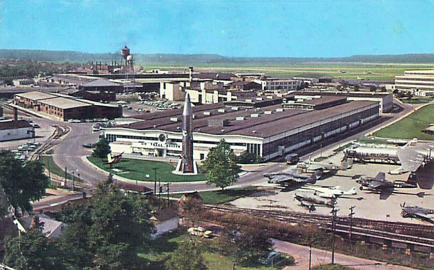 Altes Schwarz-Weiß-Foto eines Flughafens mit geparkten Flugzeugen, Gebäuden, Bäumen, Straßen, Fahrzeugen, Strommasten, Gras, Bahnschienen, Türmen, fernen Hügeln und einem klaren blauen Himmel, mit Text am unteren Rand.