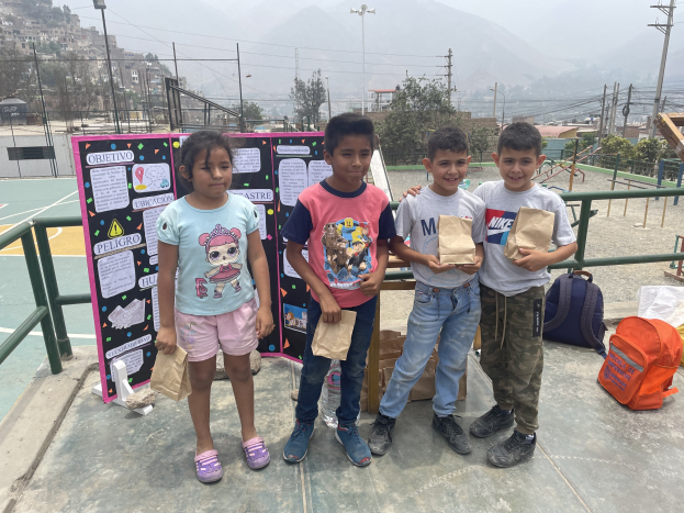 Gruppe von Kindern auf einem Basketballfeld mit Papiertüten in den Händen, Tüten und Gegenstände rechts und eine Tafel mit Text links, vor einer Außenkulisse mit Zaun, Pfosten, Gebäuden.