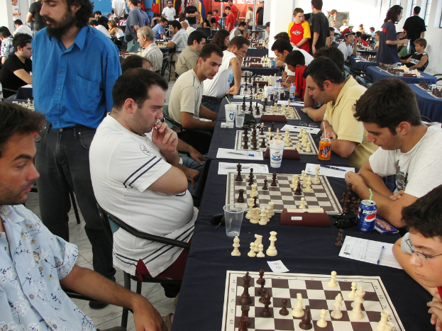 Eine Gruppe von Menschen spielt Schach mit einem Mann in einem blauen Hemd auf der linken Seite, ein Fenster in der oberen rechten Ecke und ein Tisch mit einem Glas, Schachbrett, Schachfiguren, einer Coke, Papier, Stift und einer Tischdecke.