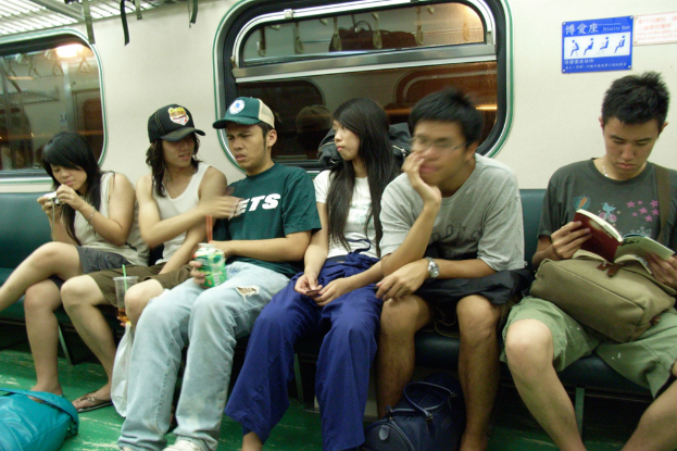 Menschen, die auf Sitzen in einer U-Bahn sitzen, wobei eine Person ein Buch hält, eine andere eine Tetra Pack, eine andere eine Dose und eine andere eine Kamera, mit Taschen auf dem Boden und Glasfenstern im Hintergrund.