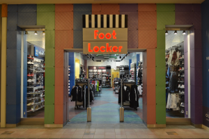 Innensicht eines Foot Locker Schuhgeschäfts mit Kleidung an Bügeln und Ständern unter Deckenbeleuchtung, mit sichtbarem Geschäftsfußboden.