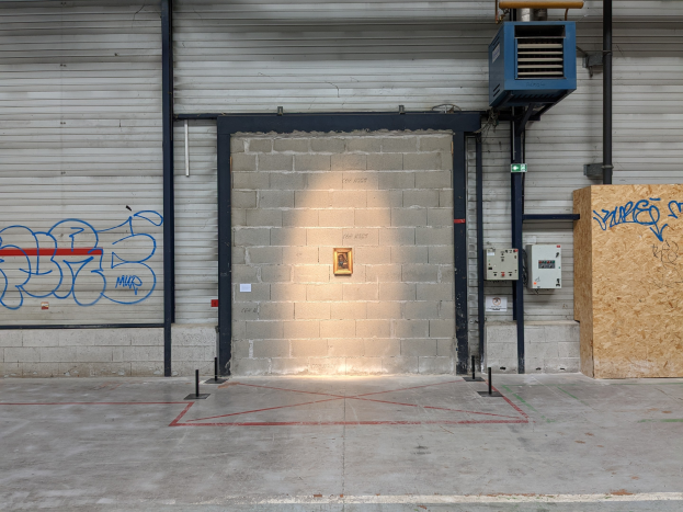 Leerer Lagerraum mit graffiti-bedeckten Wänden, einer beleuchteten Wandlampe, Metallstangen und Rollläden auf der linken Seite, einem hölzernen Objekt mit Text auf der rechten Seite und einer sichtbaren Decke.