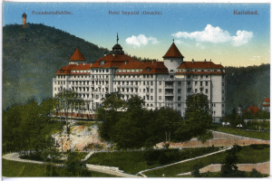 Eine alte Postkarte zeigt das Hotel Imperial in Karlsbad, Deutschland, ein großes Gebäude mit zahlreichen Fenstern, umgeben von Bäumen, Pflanzen und Gras, mit einem Weg, der zu ihm führt, vor einem Hintergrund von Hügeln unter einem bewölkten Himmel, mit Text oben auf dem Bild.