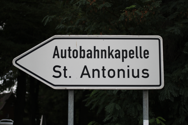 Ein weißes Schild mit der Aufschrift "Autobahnkapelle St. Antonius", das an zwei Pfosten befestigt ist, mit einem Auto auf der linken Seite und Bäumen im Hintergrund.