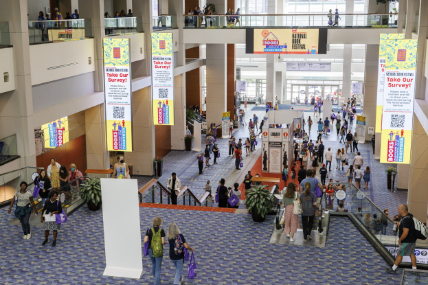 Eine große Gruppe von Menschen, die durch ein großes Gebäude mit Bannern, Topfpflanzen, Rolltreppen, Geländern, Säulen, bedruckten Schildern, Glastüren und anderen Gegenständen geht, einige tragen Taschen, wahrscheinlich beim Nationalen Buchfestival.