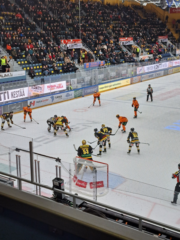 Eine Gruppe von Menschen spielt Hockey auf einem Eisstadion mit einem Netz, einer Umzäunung, sitzenden Zuschauern, Bannern und Scheinwerfern.