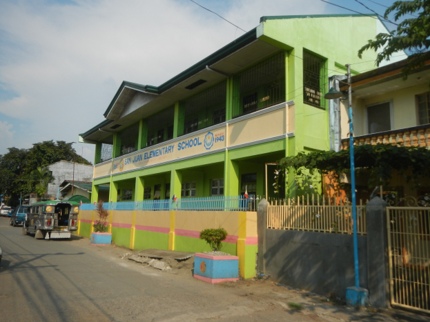 Ein Schulgebäude mit einer grünen und gelben Fassade, das sich auf einer Straße mit Fahrzeugen, Topfpflanzen, einem Zaun, einem Tor, einem Straßenpfahl, Bäumen, Drähten und einem bewölkten Himmel befindet.