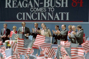 Eine Gruppe von Menschen auf einer Bühne, die klatschen, mit einem Podium und einem Mikrofon davor, Fahnen unten und eine Wand mit Text dahinter.