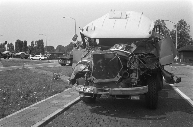 Ein Schwarz-Weiß-Foto eines schwer beschädigten Autos am Straßenrand mit eingedellter Motorhaube und zersplitterten Fenstern, während andere Fahrzeuge, Laternenpfähle, Bäume und ein Haus im Hintergrund zu sehen sind.