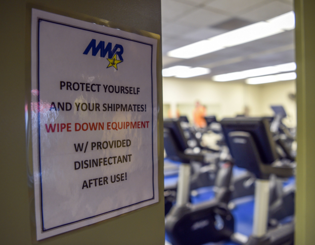 Ein Türschild mit der Aufschrift "Schützen Sie sich und Ihre Schiffskameraden" mit Laufbändern und Deckenleuchten im Hintergrund, das Bild leicht unscharf, um Bewegung anzudeuten.