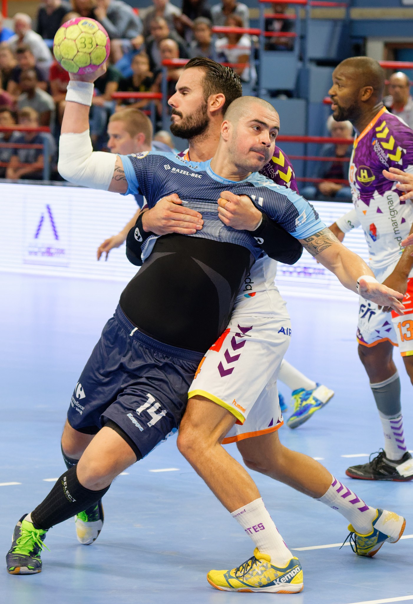 Männer beim Handballspielen in einem Stadion, einer hält den Ball, mit Zuschauern im Hintergrund.