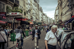 Zwei große Gebäude mit einer Straße dazwischen, wo eine Menge Menschen geht und mehrere Stände auf dem Erdgeschoss eingerichtet sind, mit einigen Mülltonnen drumherum. Ein Mann fährt ein Fahrrad auf der Straße, und Autos sind auf der gegenüberliegenden Seite geparkt.