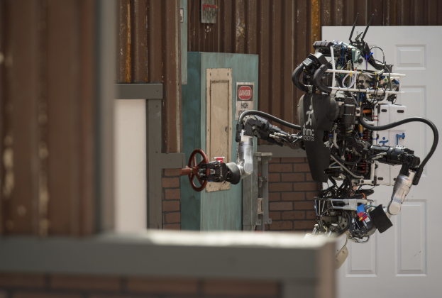 Ein humanoider Roboter mit ausgestreckten Armen und geneigtem Kopf steht vor einem Gebäude mit einer Tür und einer Wand dahinter, als würde er gleich sprechen.