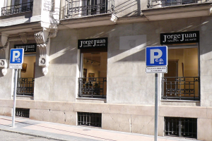 Ein mehrst├Âckiges Geb├Ąude mit Fenstern, Gel├Ąndern, Türen und S├Ąulen, das von Schildern auf Pfosten an der Stra├če und einsehbaren Innenobjekten gepr├Ągt ist, die eine belebte Stadtumgebung vermuten lassen.