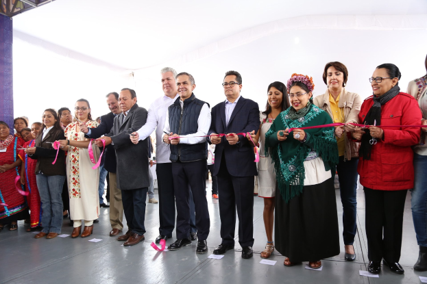 Eine Gruppe von Menschen, die eine Schnur schneiden, mit einem Pfeiler auf der linken Seite, verstreuten Papieren auf dem Boden und einem weißen Tuch im Hintergrund.
