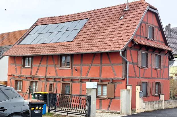 Haus mit Solarpanelen auf dem Dach, ein geparktes Auto davor, Mülltonnen, ein Tor, eine Wand, Bäume und einen Himmel im Hintergrund.