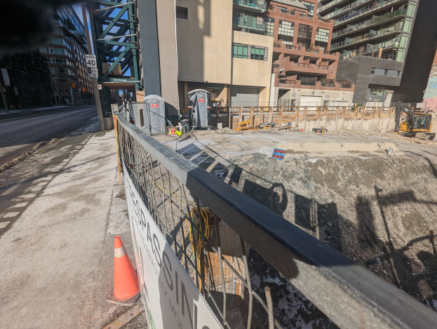 Eine Baustelle mit einem Zaun und einem Verkehrskegel im Vordergrund, eine Straße links, Gebäude im Hintergrund, ein Baumaschinenfahrzeug und ein Müllcontainer.